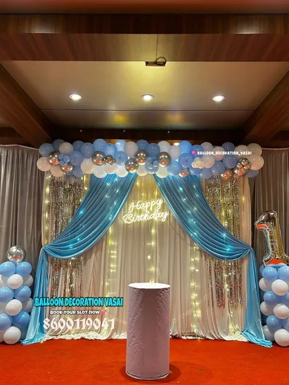 A grand stage decoration for a first birthday. The setup features blue and white drapes, a shimmer curtain with fairy lights, and a neon sign.