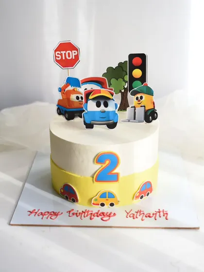 Beep beep! A fun traffic-themed cake for a second birthday. This cake is decorated with cute cartoon vehicle toppers, a stop sign, and a traffic light for a playful and colorful celebration.