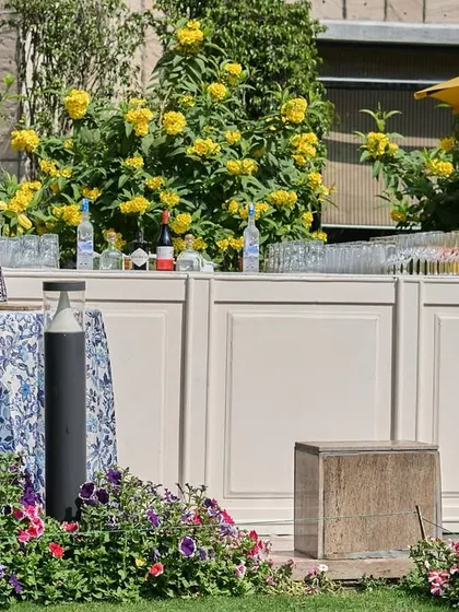 A bar setup for a garden party, with blue and white printed cocktail tables set in front of a simple white bar, backed by lush yellow-flowering bushes.