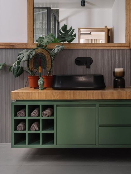 We brought a touch of nature into this Cocoon House bathroom with an olive green vanity and a wooden countertop. The design balances functionality with aesthetics, featuring open shelving for towels and a sleek black vessel sink, all complemented by a large monstera plant.