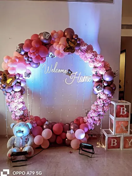 A beautiful circular balloon and floral arch in shades of pink for a welcome home party. This design includes a 'Welcome Home' neon sign, a teddy bear, and 'BABY' blocks, creating a complete and festive photo opportunity.
