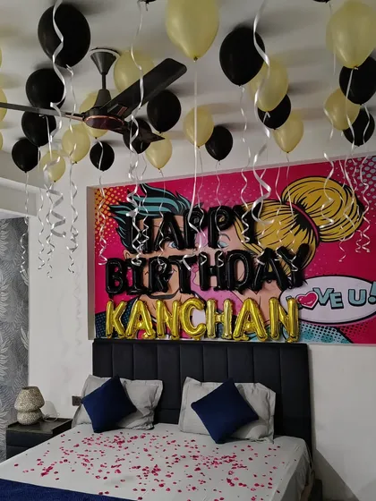A personalized birthday bedroom surprise with a pop-art backdrop. The decor features black and gold balloons, the name 'KANCHAN' in foil letters, and rose petals scattered on the bed.