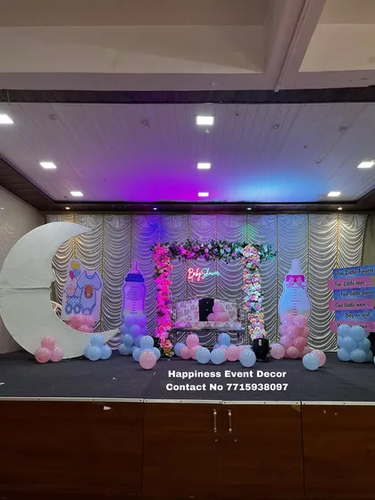 This stage setup for a baby shower features a mix of modern and playful elements. It includes a decorated swing, a 'Baby Shower' neon sign, a crescent moon prop, and large baby bottle cutouts filled with pink and blue balloons.