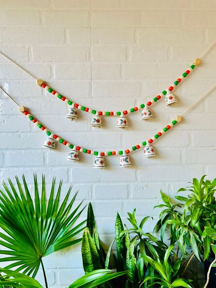 Our 'Khanak' beaded bell bunting in a festive red and green combination. This 30-inch wide bunting features five delicate mini ceramic bells and colorful wooden beads, perfect for adding charm to your entrance.