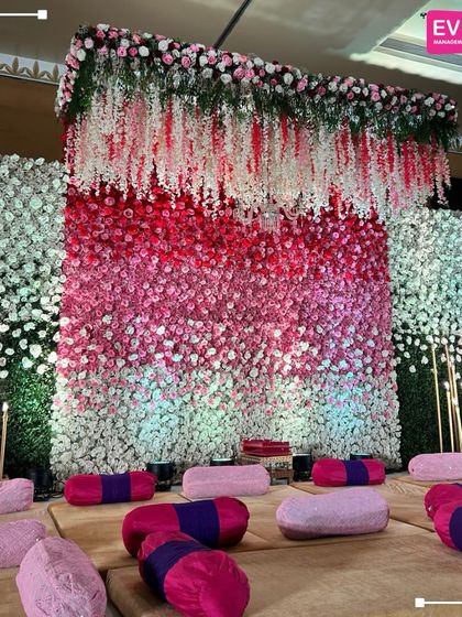 A stunning wall of flowers in an ombre pattern of pink and white serves as the backdrop for this event. The floor seating with traditional bolsters makes it a perfect setup for a Mehendi or devotional event.
