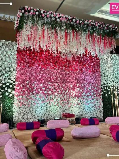 A unique and vibrant backdrop for a ceremony, featuring a wall of flowers in an ombre pattern of pink and white, with traditional seating for a fusion event.