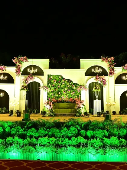 A wide view of the elegant reception stage. The green uplighting on the foliage base adds a pop of color and drama to the otherwise soft and classic white and pink decor.