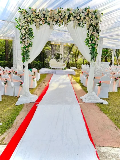 A beautiful outdoor wedding ceremony setup. The aisle is lined with elegantly decorated chairs, leading to a floral arch, all under a large white tent.