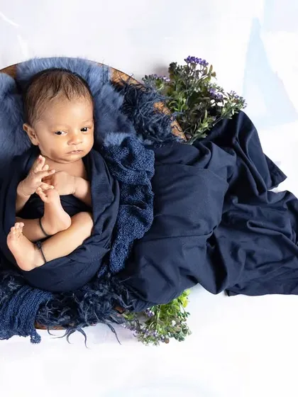 A newborn rests in a bowl, draped in a rich navy blue fabric with floral accents. The high-contrast, clean white background makes the deep colors stand out beautifully.