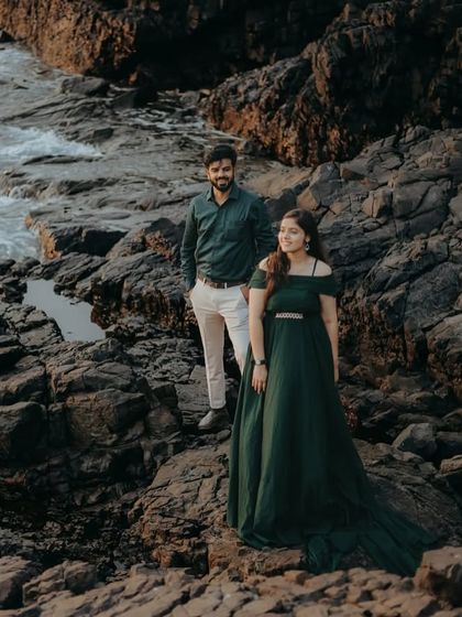 A stunning wide shot of a couple on a rocky beach. The elegant green off-shoulder gown stands out against the rugged natural landscape.