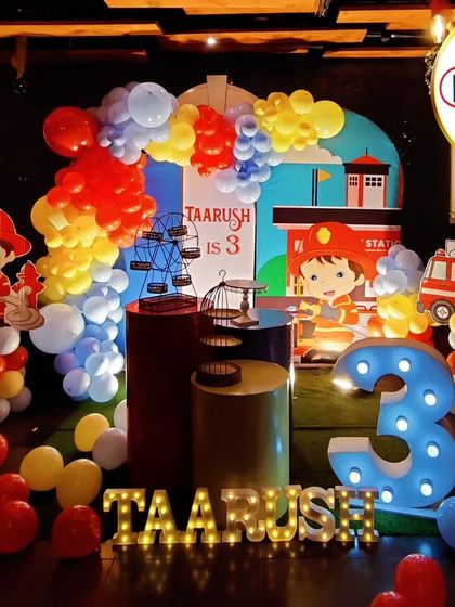 An exciting Fireman-themed third birthday party. The decor includes a fire station backdrop, firefighter character cutouts, and a large light-up number '3'.