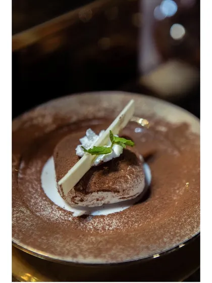 A classic Tiramisu, elegantly presented with a dusting of cocoa powder, a sprig of mint, and a white chocolate garnish. The ultimate indulgence.