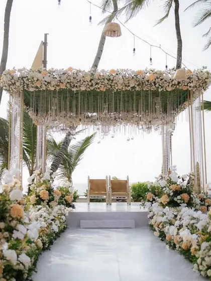 A beautiful beachside mandap setup featuring a canopy of white and peach florals with delicate crystal hangings. The design ensures the stunning sea view remains the perfect backdrop for the ceremony.