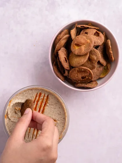 A hand dips a piece of my Bajri Wada into hummus, demonstrating the delicious combination of crunchy and creamy textures.