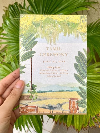 A hand holding the Tamil Ceremony invitation for a picturesque 'North meets South' wedding in a California winery. The artwork depicts a traditional South Indian ceremony setup with banana leaves and drums against a backdrop of rolling hills.