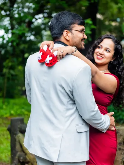 A playful and loving moment between the couple. The mom-to-be drapes a pair of tiny shoes over her partner's shoulder as they share a smile.
