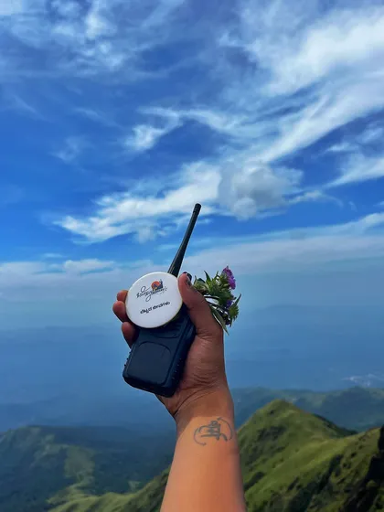A hand holding a walkie talkie with our trek badge, against the stunning backdrop of the Kumara Parvatha landscape. I use these for safety and coordination.