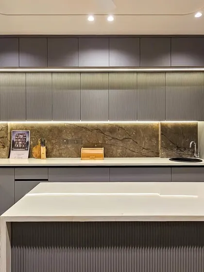 Another angle of the sleek grey kitchen, showing the island and the seamless, handle-less cabinetry. The warm lighting and natural stone backsplash prevent the grey palette from feeling cold.