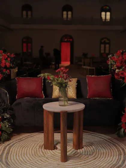 A cozy lounge seating area at the Sangeet, featuring a plush sofa with red and gold cushions, surrounded by dramatic red floral arrangements.