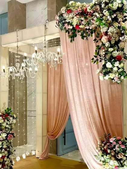 An elegant and grand entrance designed for a wedding or reception. The decor features soft pink draping, cascading floral arrangements with roses and greenery, and sparkling chandeliers that create a luxurious and welcoming ambiance for your guests.