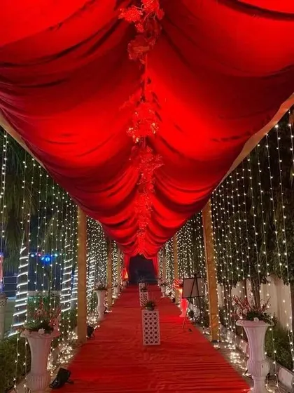 A long entrance pathway lined with trees wrapped in fairy lights. The ceiling is draped in red fabric, creating a dramatic and romantic tunnel effect for arriving guests.
