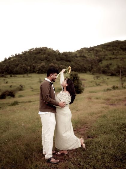 A playful and romantic wide shot of the couple in a field. She playfully touches his chin with a sunflower, a moment of lighthearted fun amidst the beauty of nature.