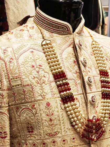 A close-up of an ivory sherwani with red and gold embroidery, paired with a traditional pearl and ruby mala. This is the epitome of groom extravagance.