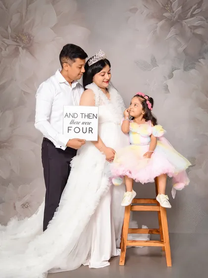 A sweet pregnancy announcement photo. The family uses a sign to share their happy news, with their daughter looking on. This classic studio portrait against a painted floral canvas is a perfect way to involve the whole family.