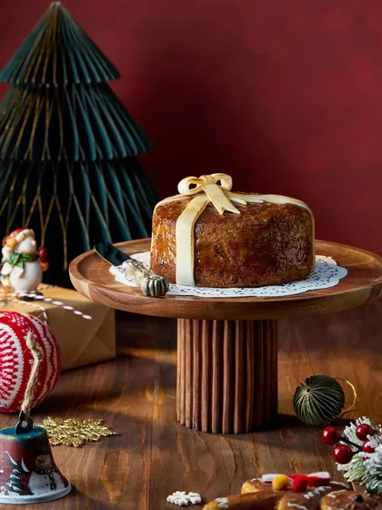 A simple yet elegant shot of a Christmas cake wrapped with a bow, placed on a wooden cake stand. The deep red and green background and festive ornaments create a rich, classic holiday image for Marriott Bonvoy.