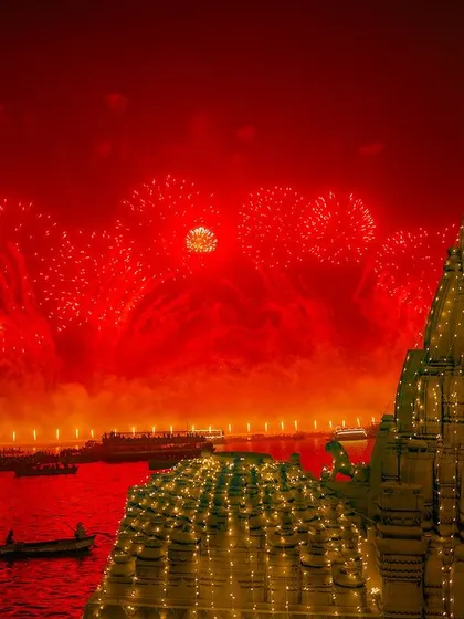 Fireworks explode in a red-tinted sky over a beautifully illuminated temple during Dev Deepawali in Varanasi. This festival of lights transforms the city into a divine spectacle.
