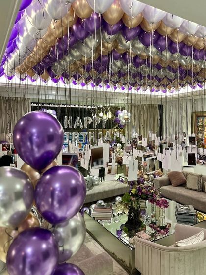 A wide view of a living room transformed for a surprise birthday party. The decor features a "Happy Birthday" balloon banner, hundreds of ceiling balloons, and personalized photo displays throughout the space.