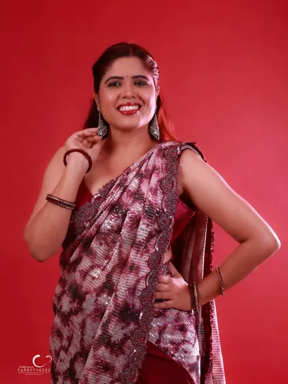 A classic pose with a hand on the hip, exuding confidence and style. The red background provides a perfect canvas for the intricate details of the saree.