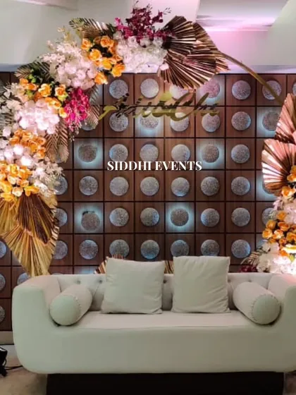 A modern and chic engagement stage with a white sofa and a backdrop of illuminated circular patterns, framed by an asymmetrical arch of lush, colorful flowers.