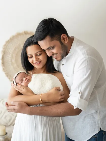 A full-length shot of new parents holding their swaddled baby girl. The coordinated white outfits create a clean and timeless look for this family portrait.
