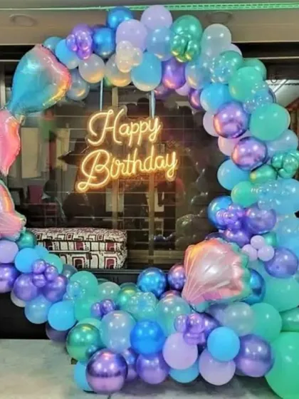 I create magical under-the-sea experiences with a mermaid-themed birthday decoration. This design features a full balloon ring in shades of teal, purple, and blue, accented with iridescent mermaid tail and seashell foil balloons. A central "Happy Birthday" neon sign adds a warm glow to the entire setup.