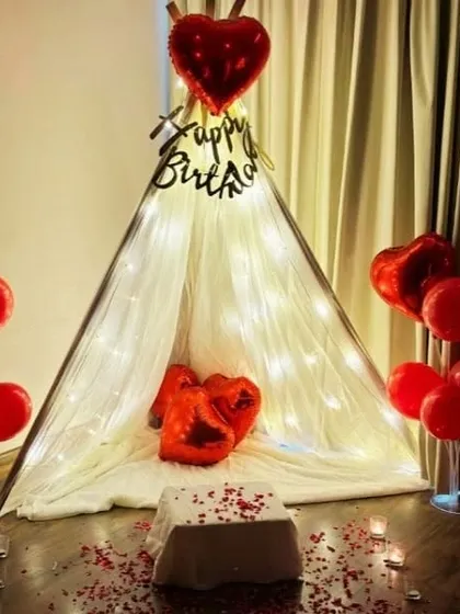 A cozy and romantic birthday surprise inside a teepee tent. This intimate setup is decorated with fairy lights, red heart-shaped foil balloons, and scattered rose petals for a truly special moment.