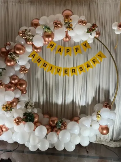 A simple yet elegant anniversary backdrop featuring a circular frame decorated with white and rose gold balloons. A "Happy Anniversary" banner is strung across the center for a personal touch.