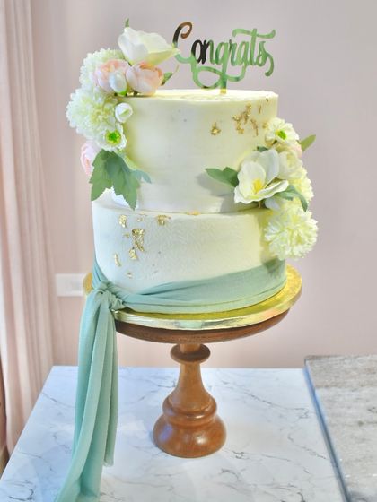 This seven-kilo, two-tier chocolate engagement cake is a statement piece. It's adorned with beautiful fresh flowers, a flowing sage green fabric drape, and delicate gold leaf for a touch of elegance.