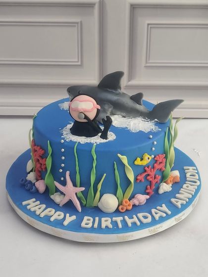A deep-sea adventure cake for a shark lover's birthday. This design features a hand-sculpted shark and scuba diver, surrounded by edible coral and seaweed on a vibrant blue fondant base.