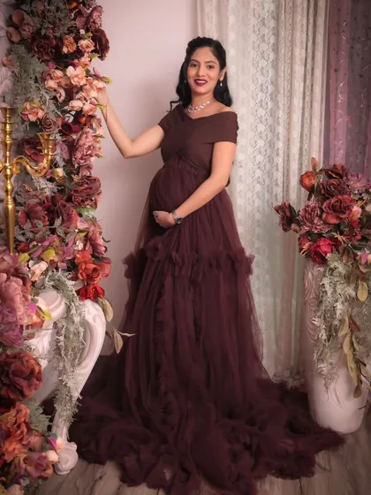 A full-length portrait showcasing the elegant off-shoulder maternity gown against our burgundy floral wall. The golden candelabra adds a classic, regal touch to the overall decor setup.