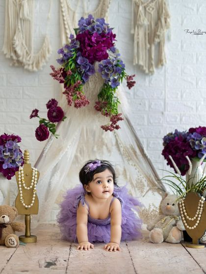 A crawling pose from a baby girl in a beautiful purple outfit. The setup with the teepee and flowers adds a touch of whimsical charm.
