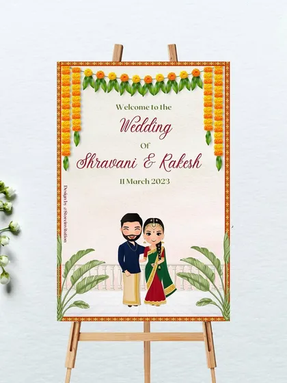This wedding welcome sign was designed for a South Indian ceremony. It features a traditional marigold toran, banana leaf illustrations, and a custom caricature of the couple in their wedding attire, offering a truly personal touch.