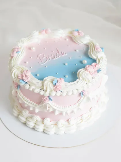 A vintage-inspired "Bride to be" cake with intricate piping and a soft pink and blue airbrushed top. This design is perfect for a bridal shower, combining classic techniques with a dreamy color palette.