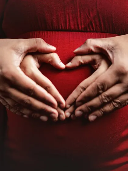 A classic symbol of love. The couple's hands form a heart shape over the baby bump, a simple yet powerful statement of their love for their unborn child, highlighted by the vibrant red dress.