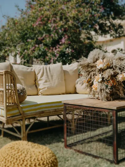 A sun-drenched lounge corner. This setup features a comfortable cane sofa, a knitted yellow pouf, and a rustic industrial-style coffee table, perfect for a relaxed daytime event.