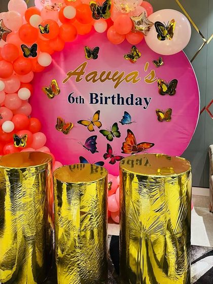 A vibrant butterfly theme for a 6th birthday. The pink balloon arch and backdrop are covered in colorful butterflies, with golden pedestals for the cake.