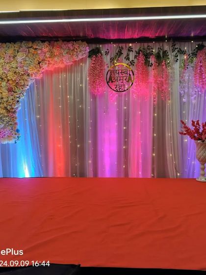 A stage backdrop for a Parna ceremony, featuring a floral corner arrangement, hanging flowers, and a fairy light curtain with colorful uplighting.