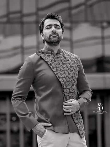 A black and white portrait of Abir Chatterjee against a modern architectural background. The strong lines of the building complement the sharp tailoring of his outfit.