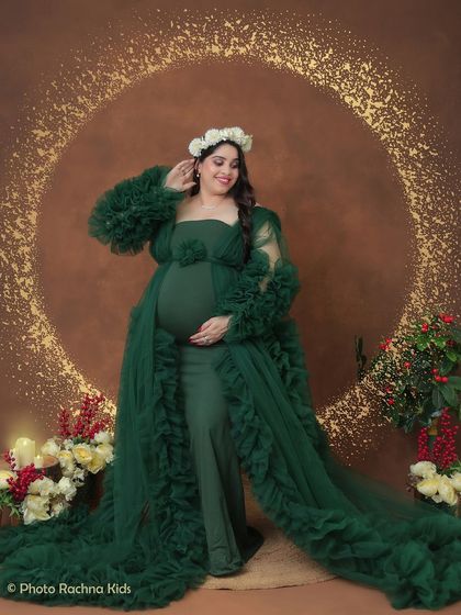 A smiling portrait against a glittering gold backdrop. The mom-to-be looks radiant in a green ruffled gown, complete with a delicate floral crown.