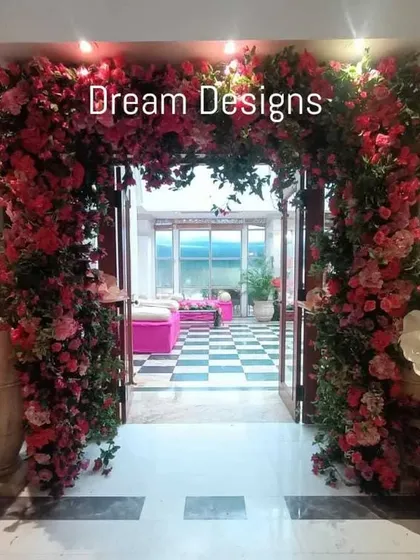 A grand floral entrance designed for a Mehendi party. The doorway is framed by a massive arch of pink flowers and oversized paper blooms, creating a vibrant and joyful welcome.